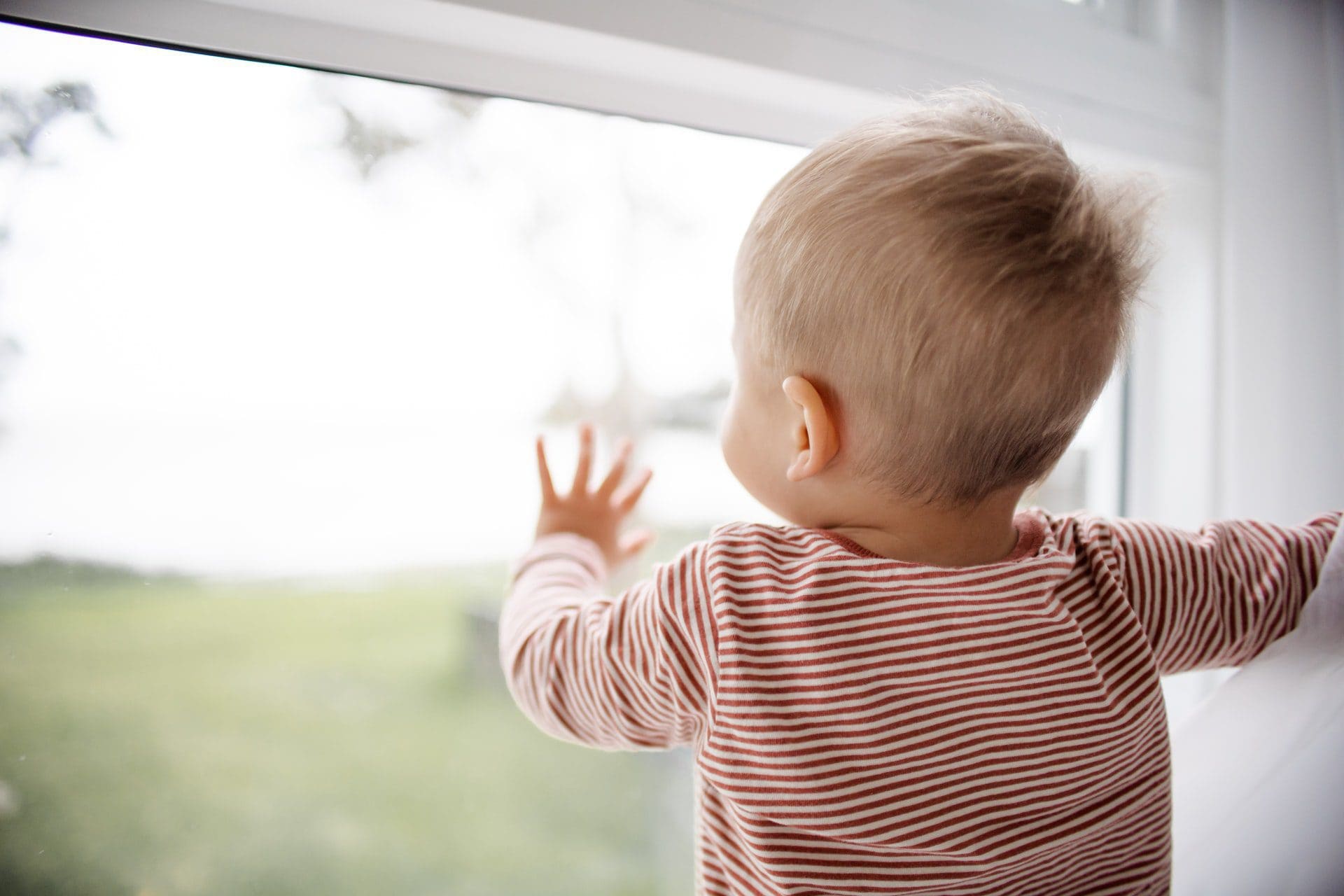 A toddler with blond hair, wearing a red and white stripped shirt, looks outside through glass while touching the glass.
